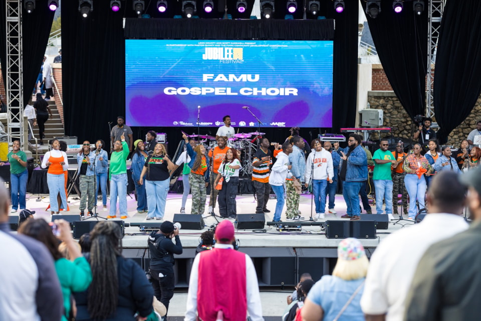 The FAMU Gospel Choir performs energetically on stage at the Jubilee Gospel Music Festival, wearing green and orange attire in front of a large digital screen.