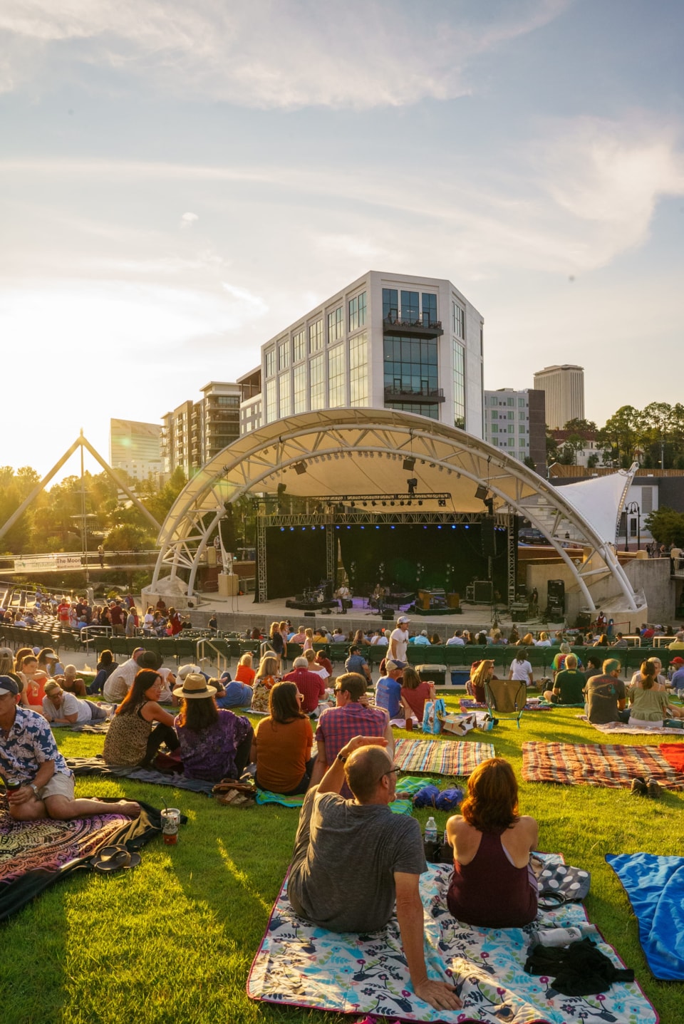 Concertgoers relax on blankets across the sunlit grassy lawn, watching a live performance beneath the canopy of the Adderley Amphitheater.