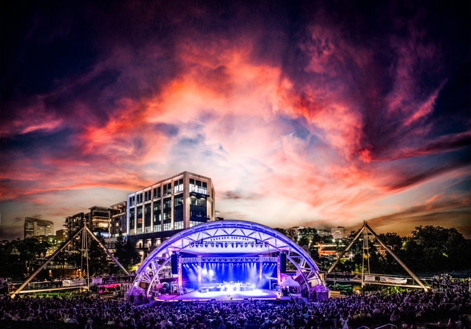 A vibrant sunset of orange, pink, and purple clouds frames the packed Adderley Amphitheater as a crowd watches Travis Tritt perform under blue stage lighting.