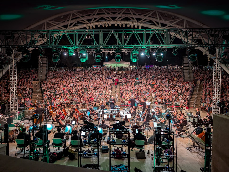 The Tallahassee Symphony Orchestra performs 'A John Williams Cinematic Celebration' under green stage lights, while a massive, packed crowd fills the tiered seating at the Adderley Amphitheater.
